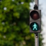 Green pedestrian signal lit, indicating walk, with trees and foliage in bright daylight.