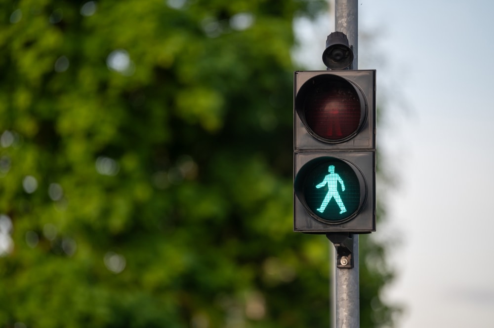 Green pedestrian signal lit, indicating walk, with trees and foliage in bright daylight.