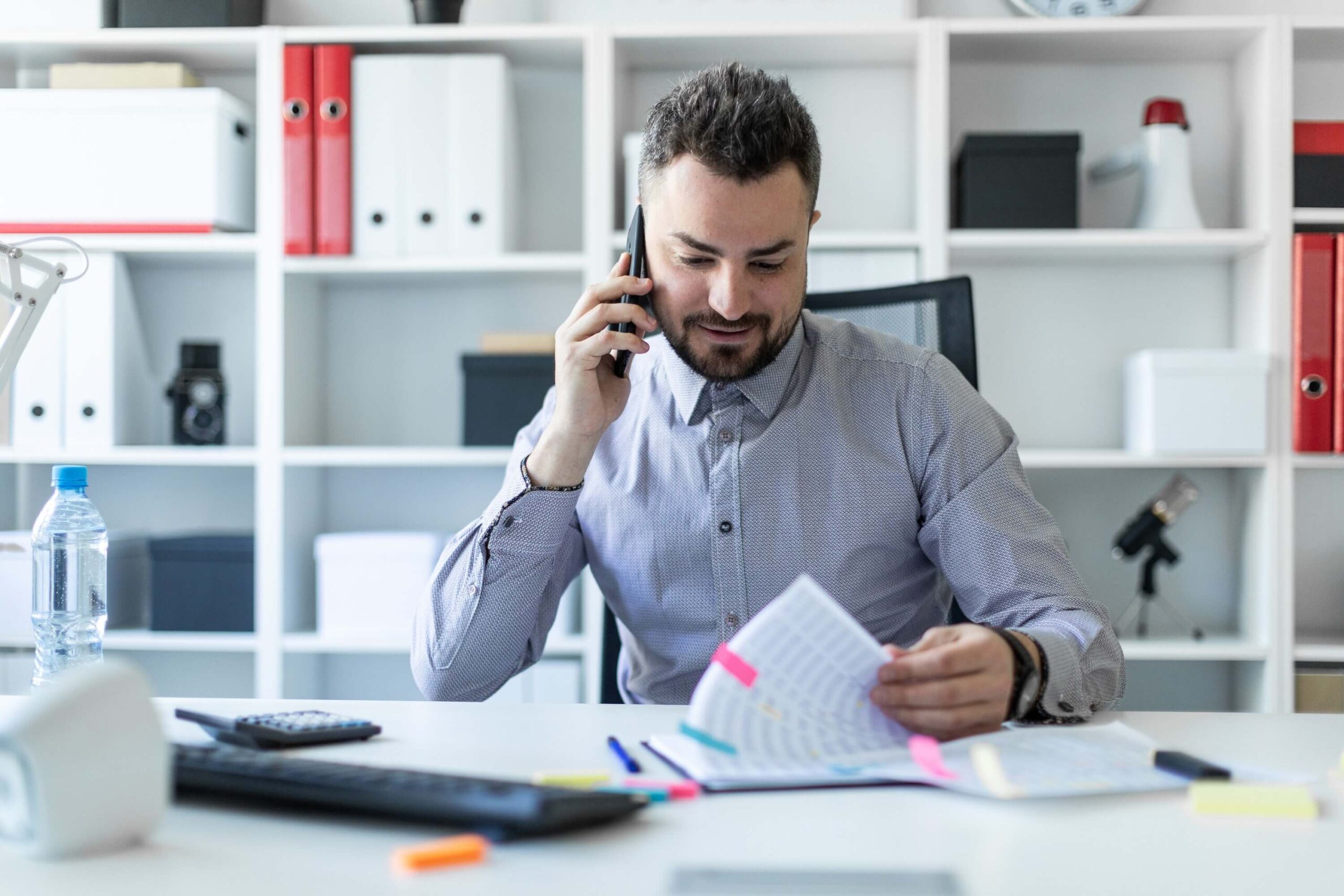 A young man is sitting in the office, talking on the phone and working with documents.