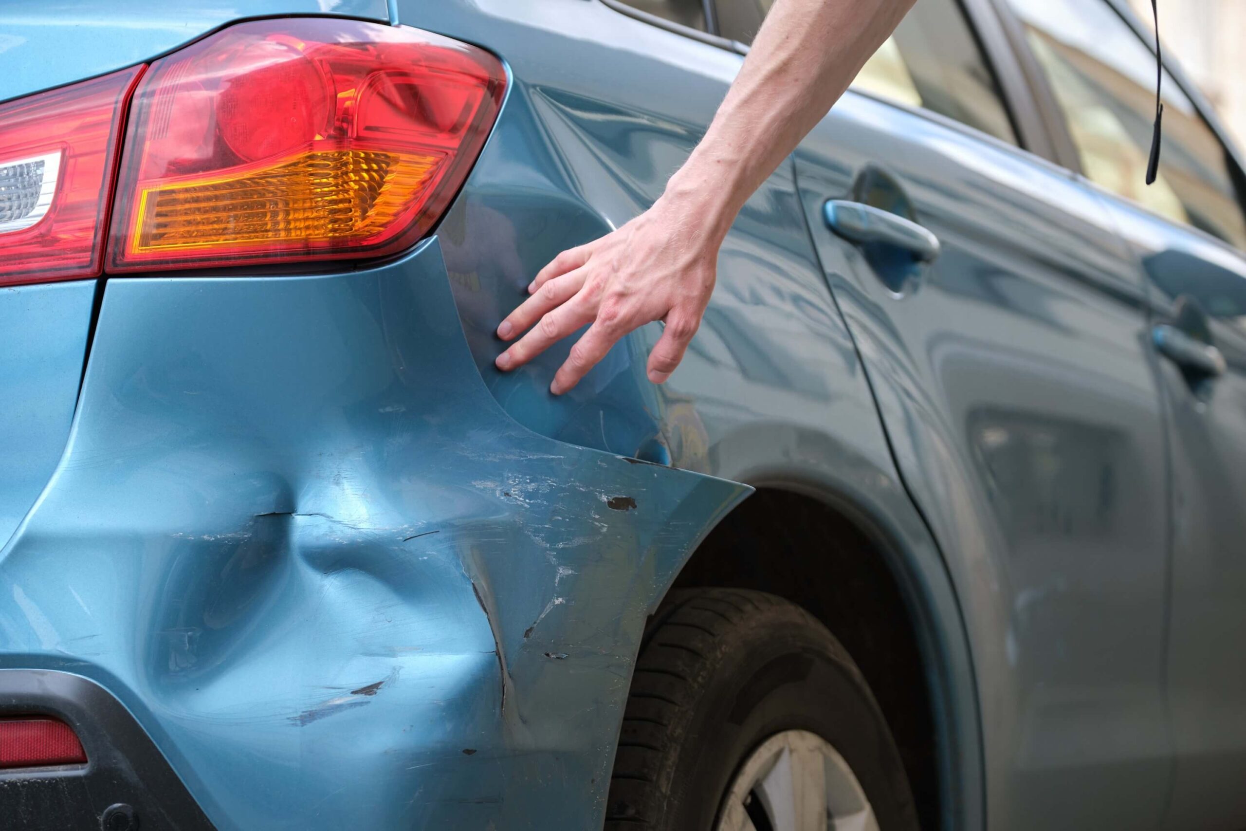 Hand of driver examining car with damaged fender parked on side of city street