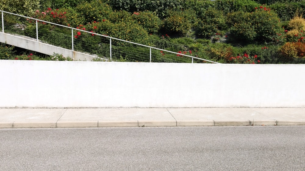 White plaster fence with flowering garden and  a staircase on behind. Cement sidewalk and street in front. Background for copy space.