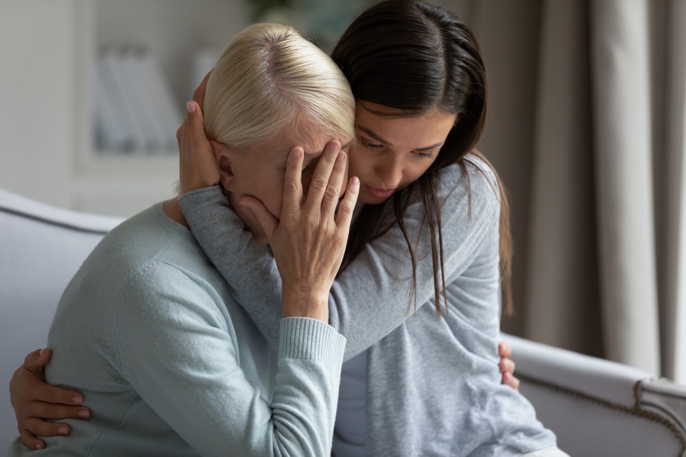  Empathic young lady embracing soothing crying depressed elder mommy, sitting together at home. Upset loving grown up millennial daughter cuddling supporting depressed stressed middle aged mother.